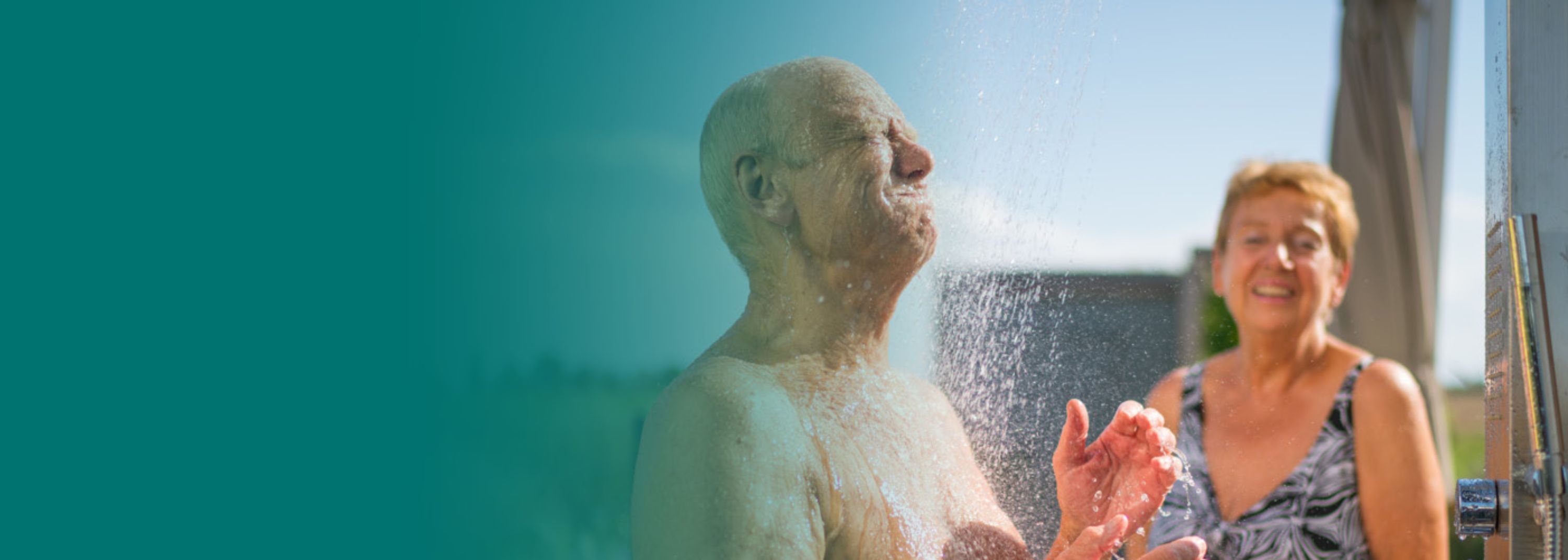 Älterer Mann erfrischt sich unter einer Außendusche, Gesicht nach oben gereckt, Wasser läuft über seinen Kopf. Im Hintergrund lächelt eine Frau mittleren Alters. Sonniges Sommerwetter.